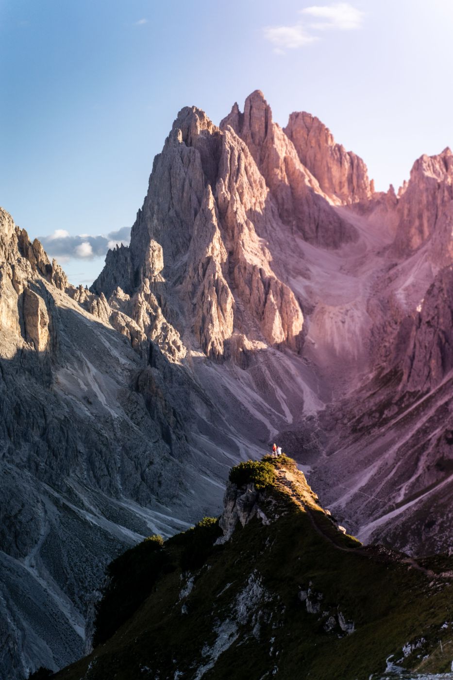Two tiny figures on a green ridge below vast Dolomite peaks lit rose-gold by the low sun
