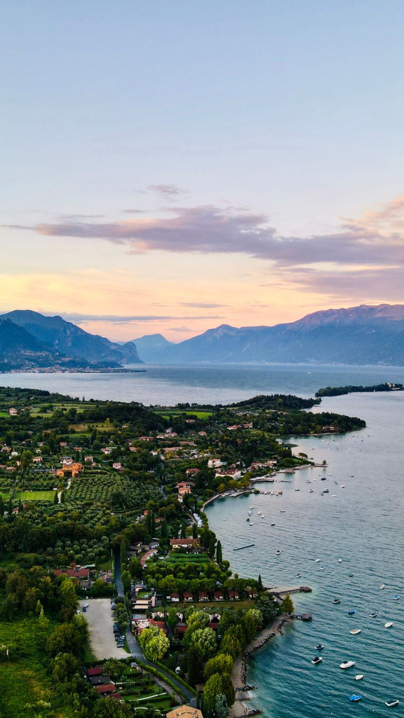 Lake Garda at dusk seen from above — olive terraces and cypress running down to the water, mountains fading pink beyond
