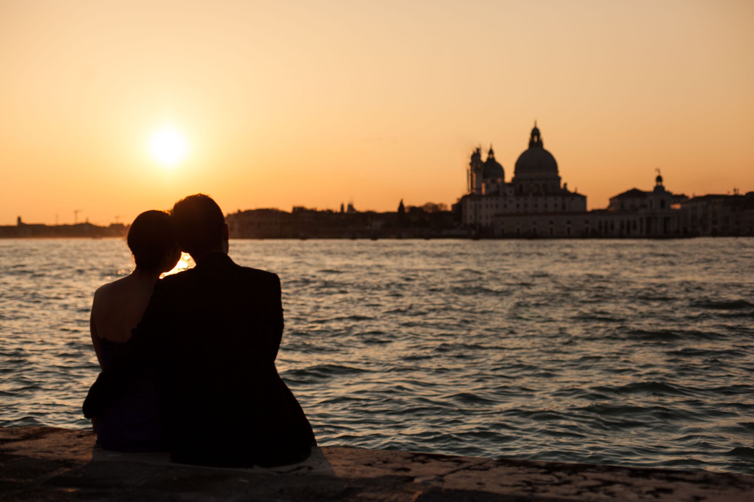 A couple sitting close together in silhouette at sunset, watching the water with a domed basilica on the horizon